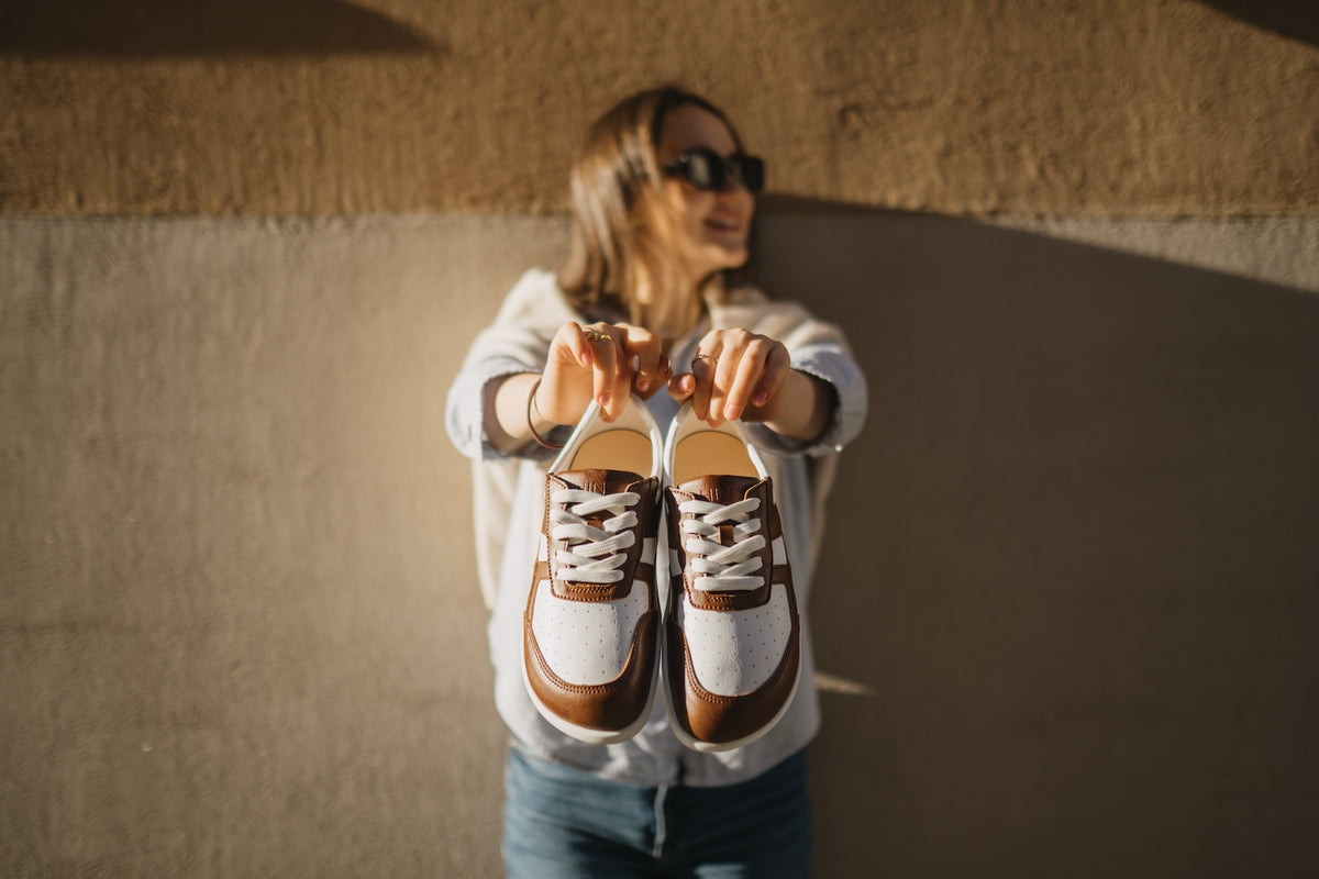 Person holding two pairs of brown and white shoes against a textured wall.